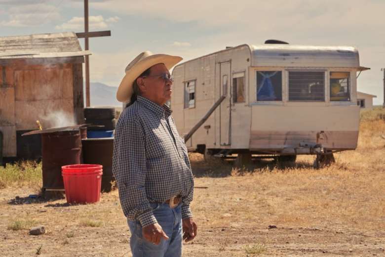 A photograph of tribal elder Arnold Sam standing outside in front of a camper and makeshift wooden building