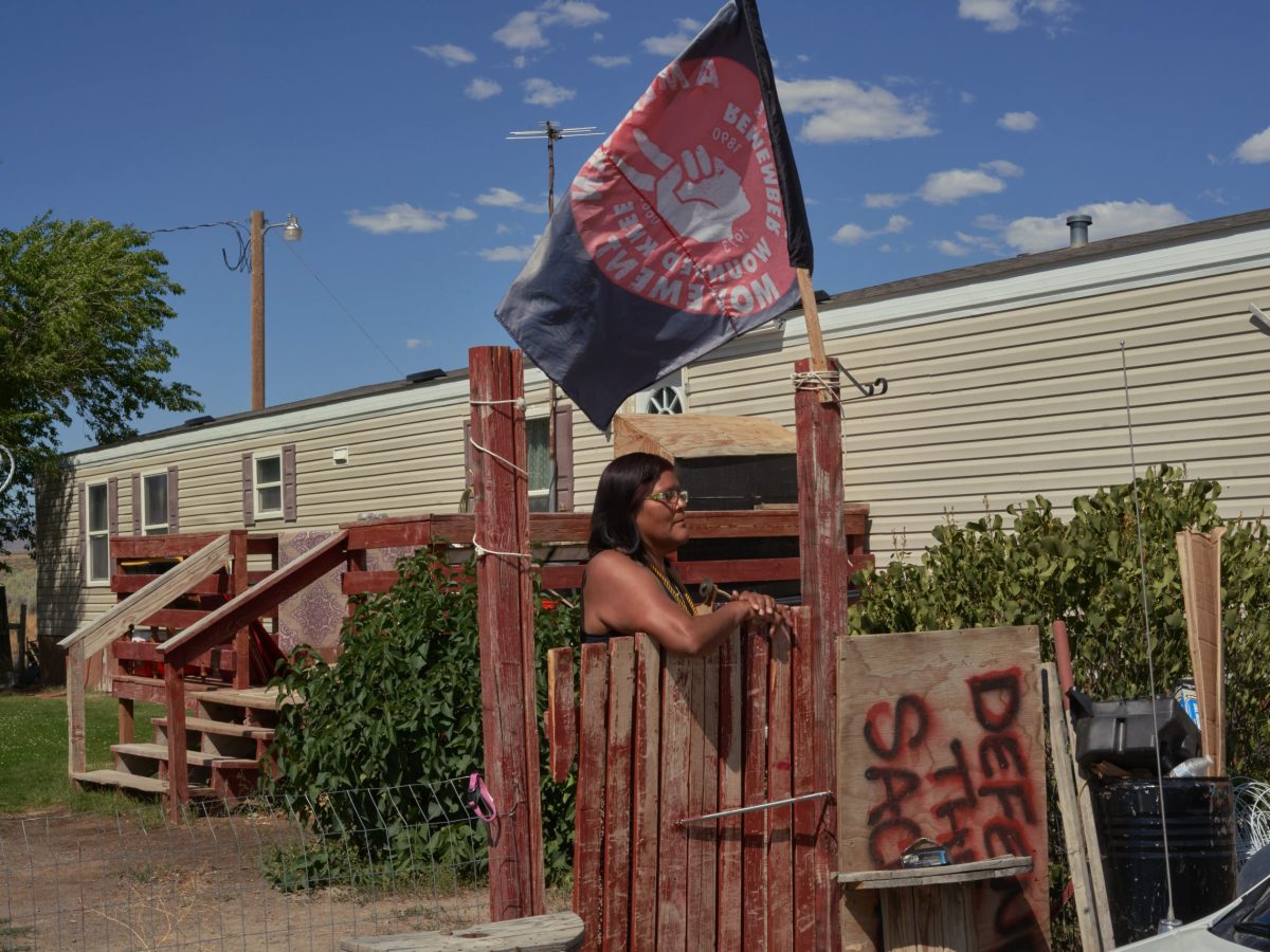 Dorece Sam stands at a red, faded gate in front of a tan mobile home. Above her flies a red and black flag that compels viewers to remember Wounded Knee.