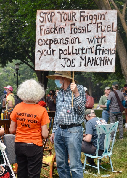 A masked protester holds a sign, which reads, "Stop you friggin' frackin' fossil fuel expansion with your pollutin' friend Joe Manchin." A second protester, facing away from the camera, wears a bright orange shirt, the back of which reads, "Protect what you love."