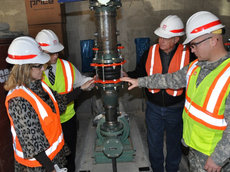 Four people in hard hats and neon reflective vests observe a metal device that is a ground-source heat pump.