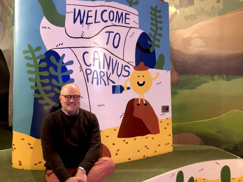A man in glasses and a sweater sits on a giant bench painted with the words "Welcome to Canvus Park"