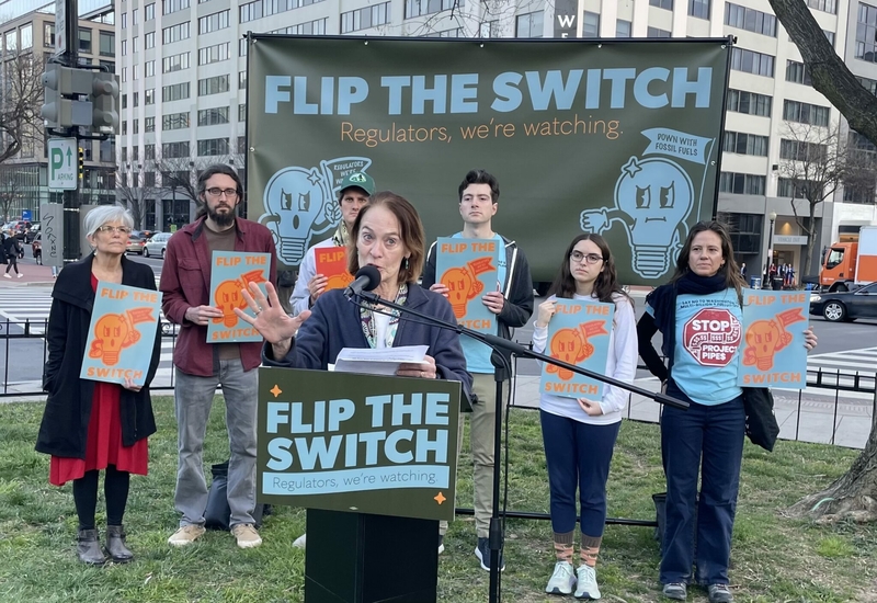 A woman speaks behind a lectern with a sign that says "flip the switch: regulators, we're watching."