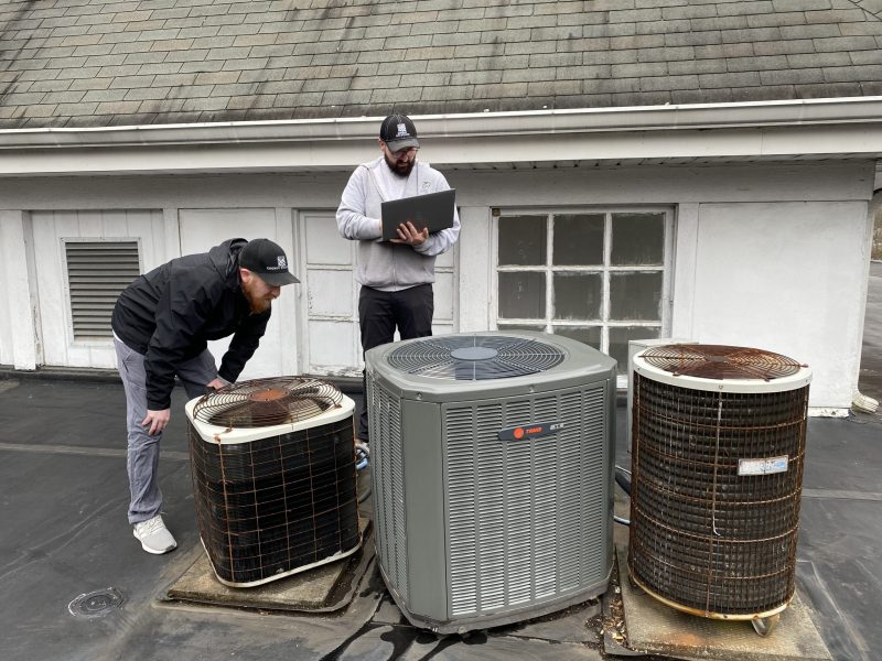 Two men stand around three old air conditioning units, with one bending to examine a unit closely and the other taking notes on a laptop.
